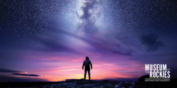 A person silhouetted on rocky ground gazing at a vibrant sunset and the Milky Way across a starry sky, with venue branding bottom right.