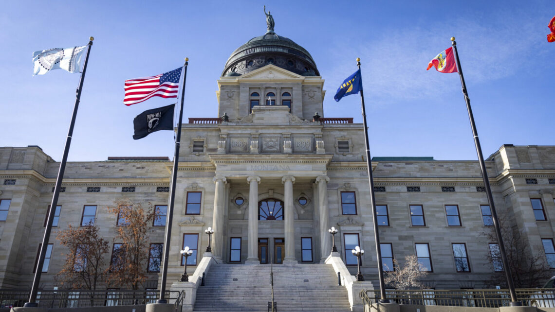 Montana Capitol building with a domed roof, grand columns, and flagpoles flying US and state flags in front of a stone facade.