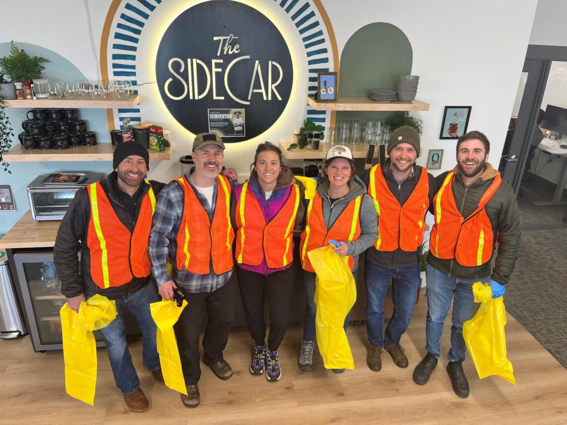 Six volunteers in orange safety vests pose for a photo inside a cafe, with a large illuminated sign reading 'The Sidecar' behind them and yellow bags in hand.