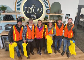 Six volunteers in orange safety vests pose for a photo inside a cafe, with a large illuminated sign reading 'The Sidecar' behind them and yellow bags in hand.