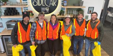Six volunteers in orange safety vests pose for a photo inside a cafe, with a large illuminated sign reading 'The Sidecar' behind them and yellow bags in hand.