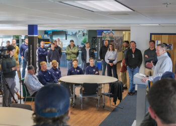 Press conference in a university lobby: athletes in blue shirts sit around a round table as a speaker at a podium addresses the crowd, with a camera crew recording on the left.