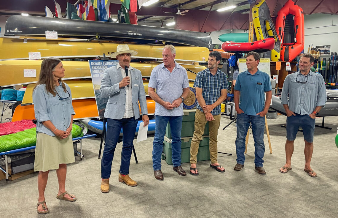 Six adults stand in a boat shop showroom; a man in a light blazer speaks into a microphone while others listen, with kayaks and gear stacked behind them.