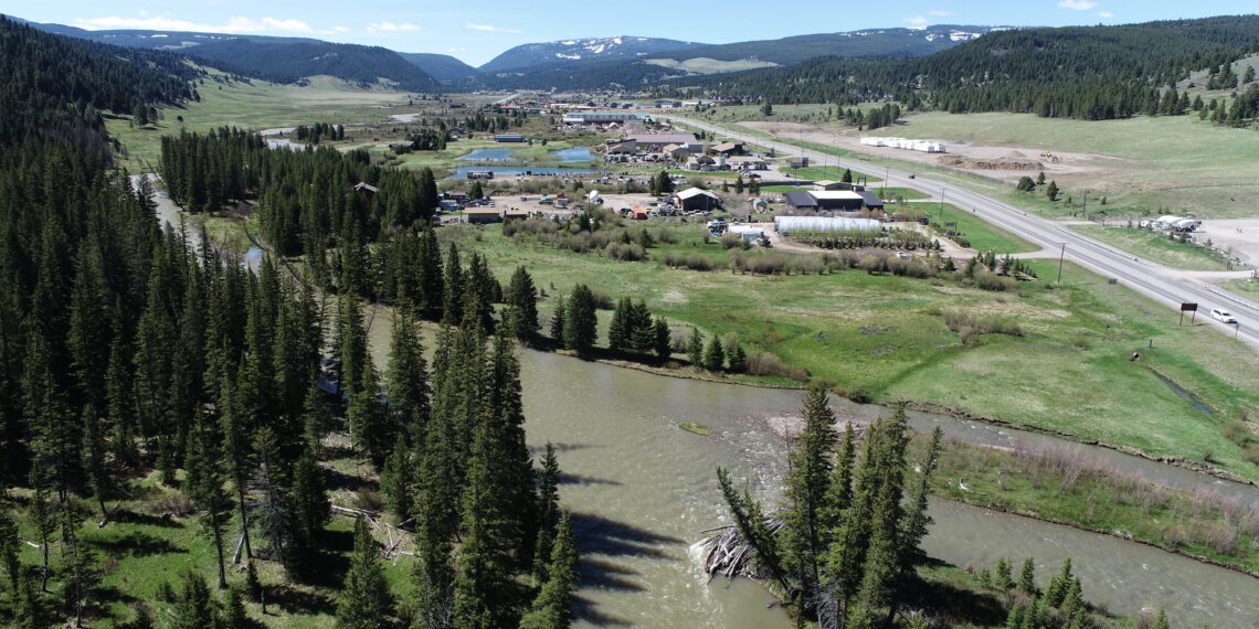 Aerial view of a small town in a green valley, with a winding river, fields, and forested hills under a blue sky.