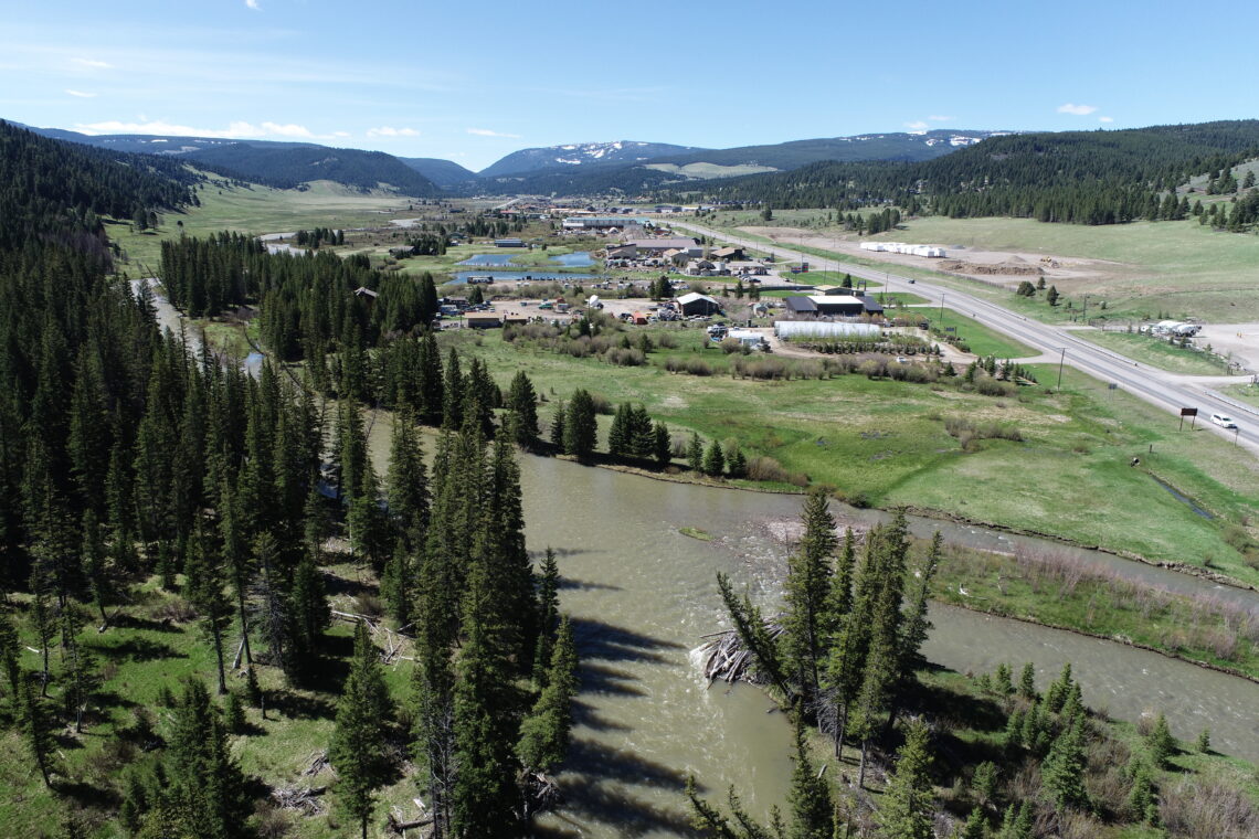 Aerial view of a small town in a green valley, with a winding river, fields, and forested hills under a blue sky.