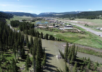 Aerial view of a small town in a green valley, with a winding river, fields, and forested hills under a blue sky.
