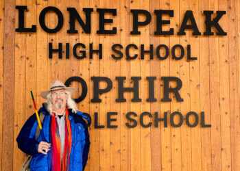 Man with hat and colorful scarf posing in front of a wooden wall that reads 'LONE PEAK HIGH SCHOOL'.