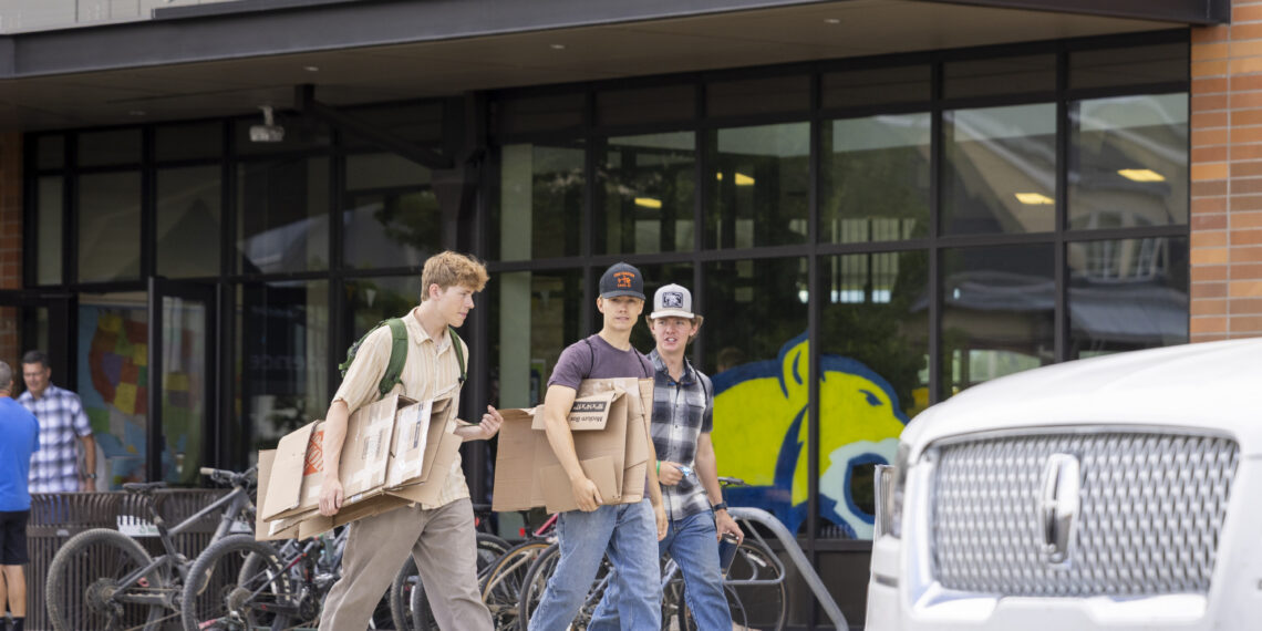 Three young men carrying large cardboard boxes outside Yellowstone Hall on a sidewalk.