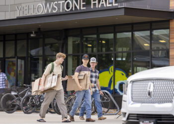 Three young men carrying large cardboard boxes outside Yellowstone Hall on a sidewalk.