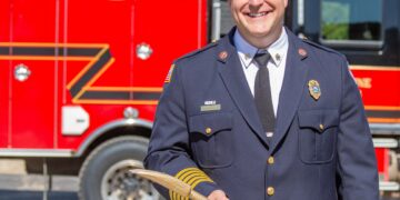 Firefighter in dress uniform holding a wooden tool, standing in front of a red fire truck with 'BIG SKY FIRE' lettering and a forested, mountainous backdrop.