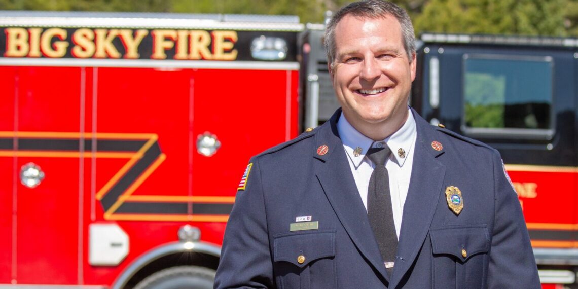 Firefighter in dress uniform holding a wooden tool, standing in front of a red fire truck with 'BIG SKY FIRE' lettering and a forested, mountainous backdrop.
