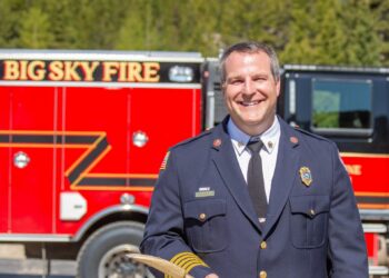 Firefighter in dress uniform holding a wooden tool, standing in front of a red fire truck with 'BIG SKY FIRE' lettering and a forested, mountainous backdrop.