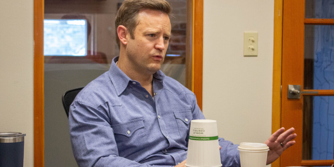 Man in a blue shirt seated at a conference table, gesturing while talking, with stacked white cups in the foreground.