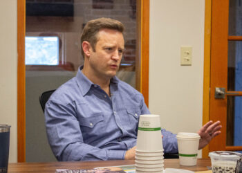 Man in a blue shirt seated at a conference table, gesturing while talking, with stacked white cups in the foreground.