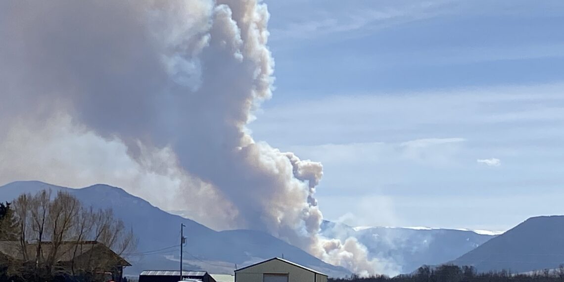 Large white smoke plume rising from a distant hillside behind a small town, with houses and vehicles in the foreground.