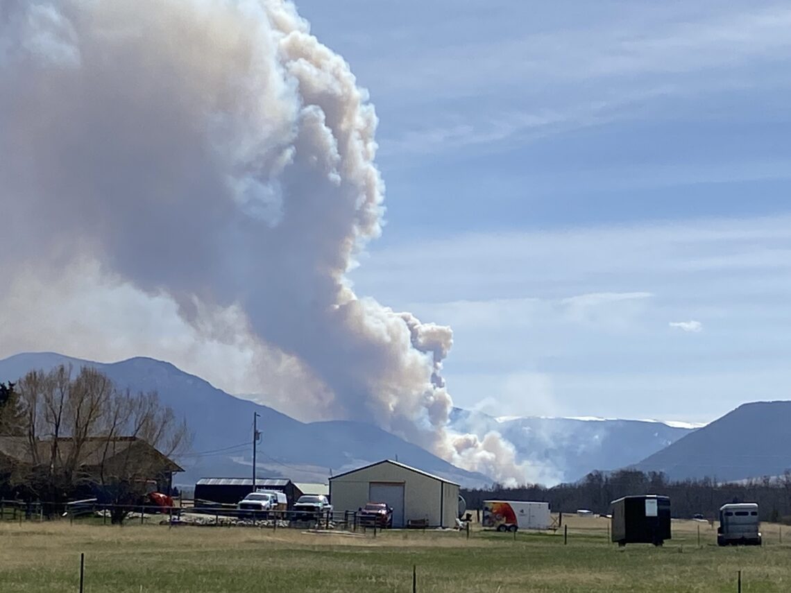 Large white smoke plume rising from a distant hillside behind a small town, with houses and vehicles in the foreground.