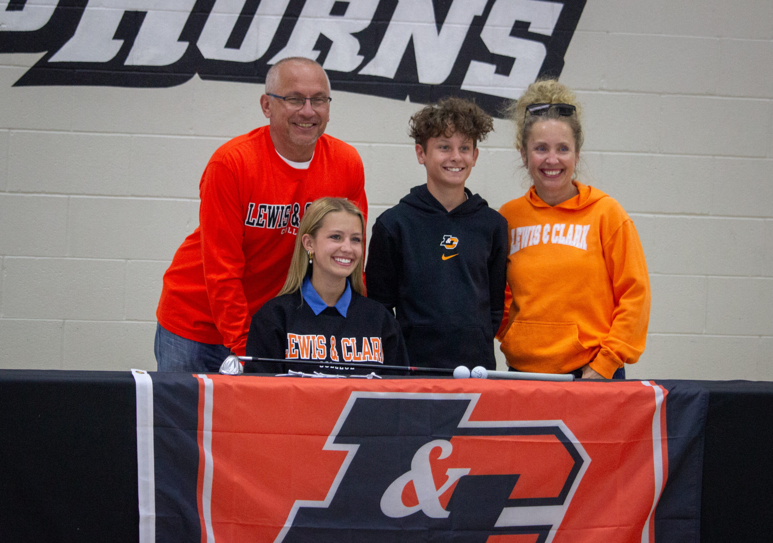 Four people stand behind a table with a large orange/navy L&C banner, wearing Lewis & Clark apparel.