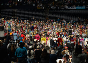 Crowd of dancers in colorful regalia performing at a powwow inside a large arena, with spectators in the stands behind them.
