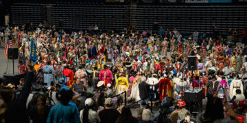 Crowd of dancers in colorful regalia performing at a powwow inside a large arena, with spectators in the stands behind them.