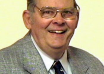 Smiling older man in a gray plaid suit, white shirt, and striped tie, wearing glasses.
