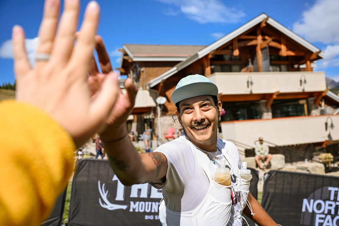 Smiling man in a light blue cap selfies at an outdoor race finish, with a blurred hand in the foreground and a wooden building behind.