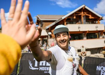 Smiling man in a light blue cap selfies at an outdoor race finish, with a blurred hand in the foreground and a wooden building behind.