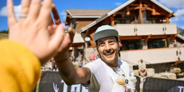 Smiling man in a light blue cap selfies at an outdoor race finish, with a blurred hand in the foreground and a wooden building behind.