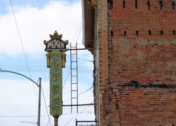 Vertical ornamental sign reading 'PEKIN(G)' attached to a building, with a brick wall and clear blue sky behind.