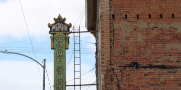 Vertical ornamental sign reading 'PEKIN(G)' attached to a building, with a brick wall and clear blue sky behind.