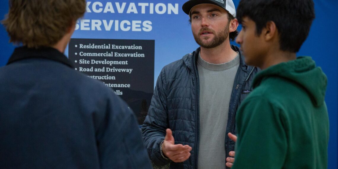 Man with a beard in a gray shirt and cap explains something to two others in front of a blue sign that says “EXCAVATION SERVICES.”