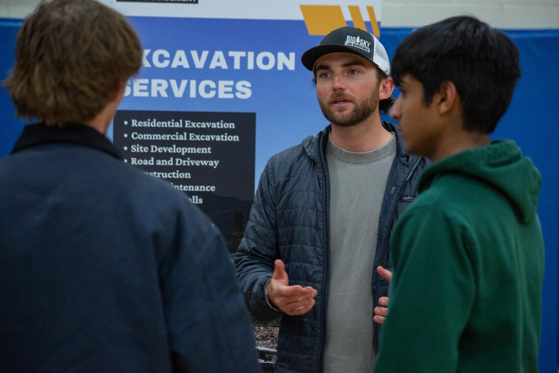 Man with a beard in a gray shirt and cap explains something to two others in front of a blue sign that says “EXCAVATION SERVICES.”