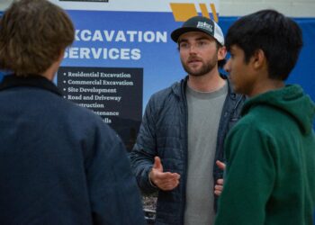Man with a beard in a gray shirt and cap explains something to two others in front of a blue sign that says “EXCAVATION SERVICES.”