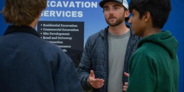 Man with a beard in a gray shirt and cap explains something to two others in front of a blue sign that says “EXCAVATION SERVICES.”