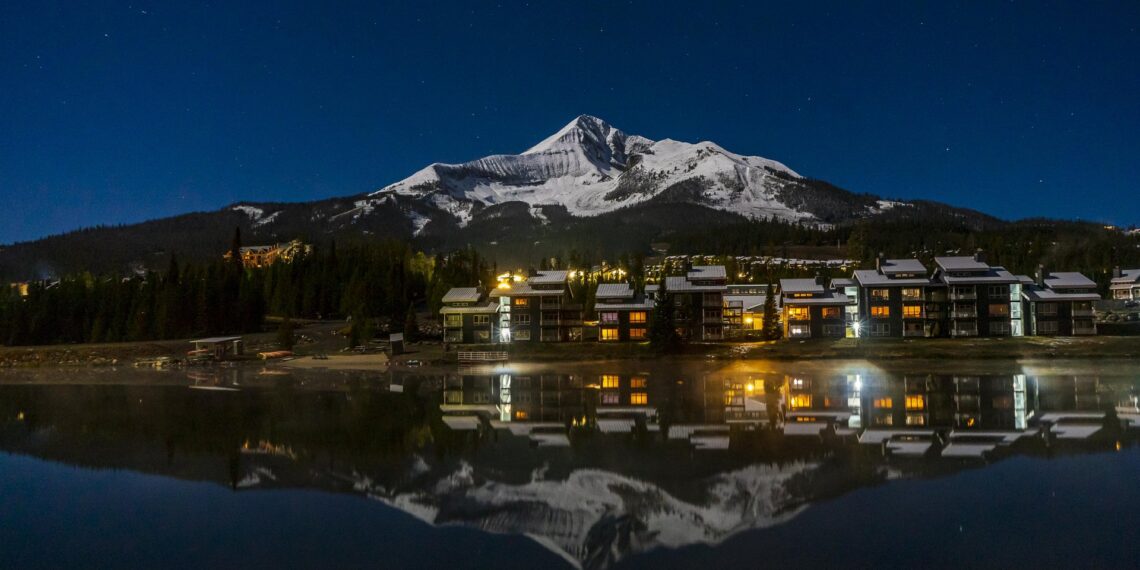 Night view of a snow-capped mountain behind a lakeside town with lit windows, reflected in calm water at dusk/nightfall.