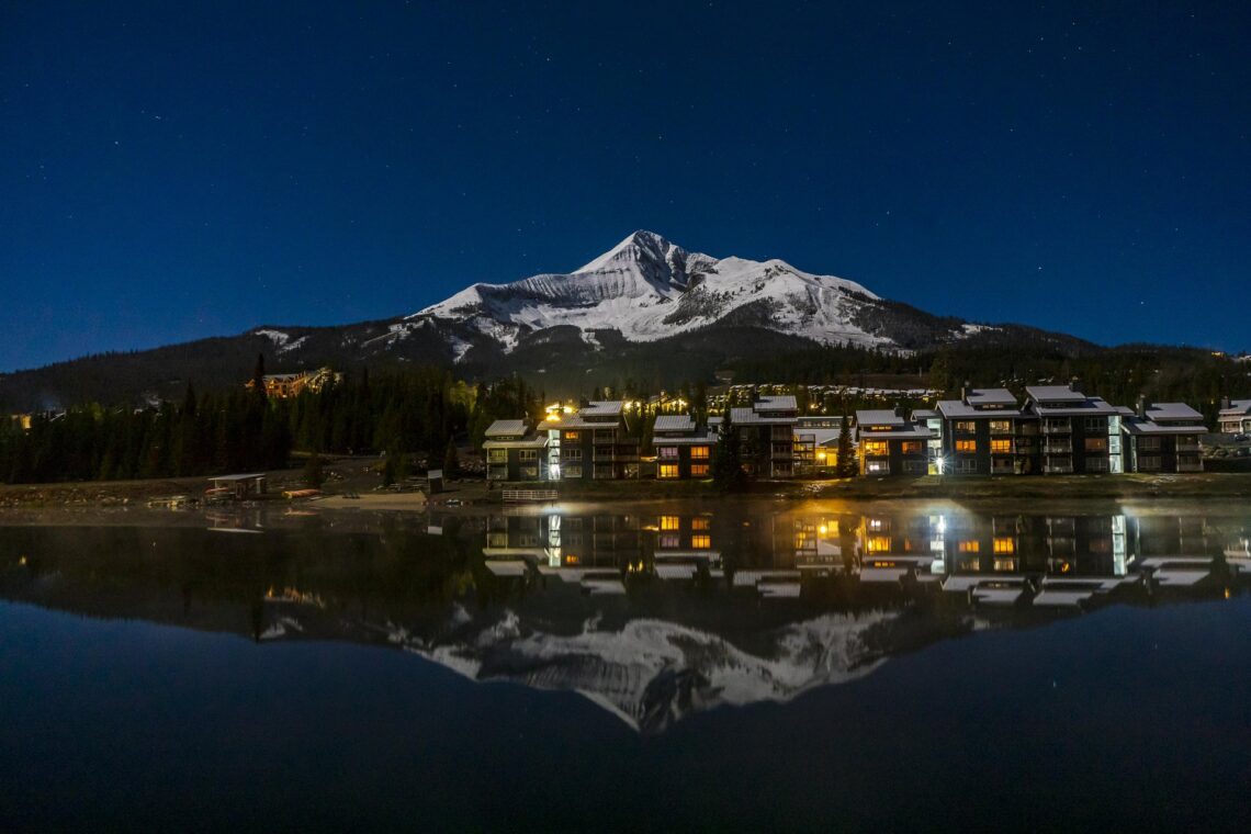 Night view of a snow-capped mountain behind a lakeside town with lit windows, reflected in calm water at dusk/nightfall.