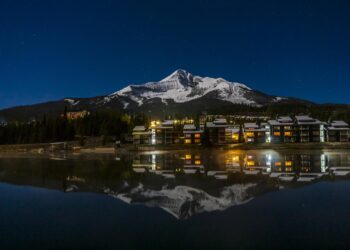 Night view of a snow-capped mountain behind a lakeside town with lit windows, reflected in calm water at dusk/nightfall.