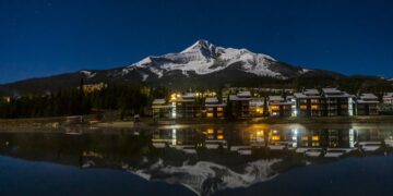 Night view of a snow-capped mountain behind a lakeside town with lit windows, reflected in calm water at dusk/nightfall.