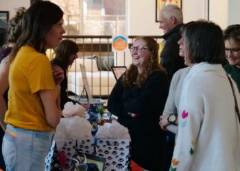 Group of people chat at a booth with gift bags and brochures in a bright indoor venue, smiling and engaged in conversation.