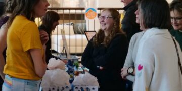 Group of people chat at a booth with gift bags and brochures in a bright indoor venue, smiling and engaged in conversation.