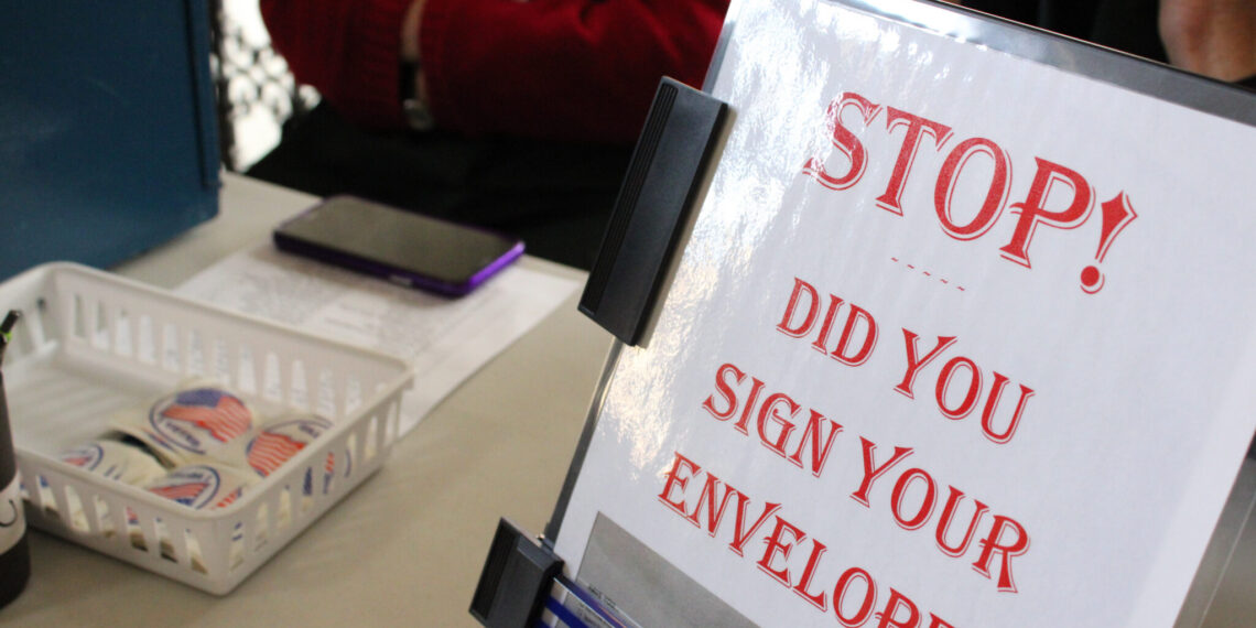 Signage on a desk asking: 'STOP! Did you sign your envelope?' with a binder and basket of stickers nearby.
