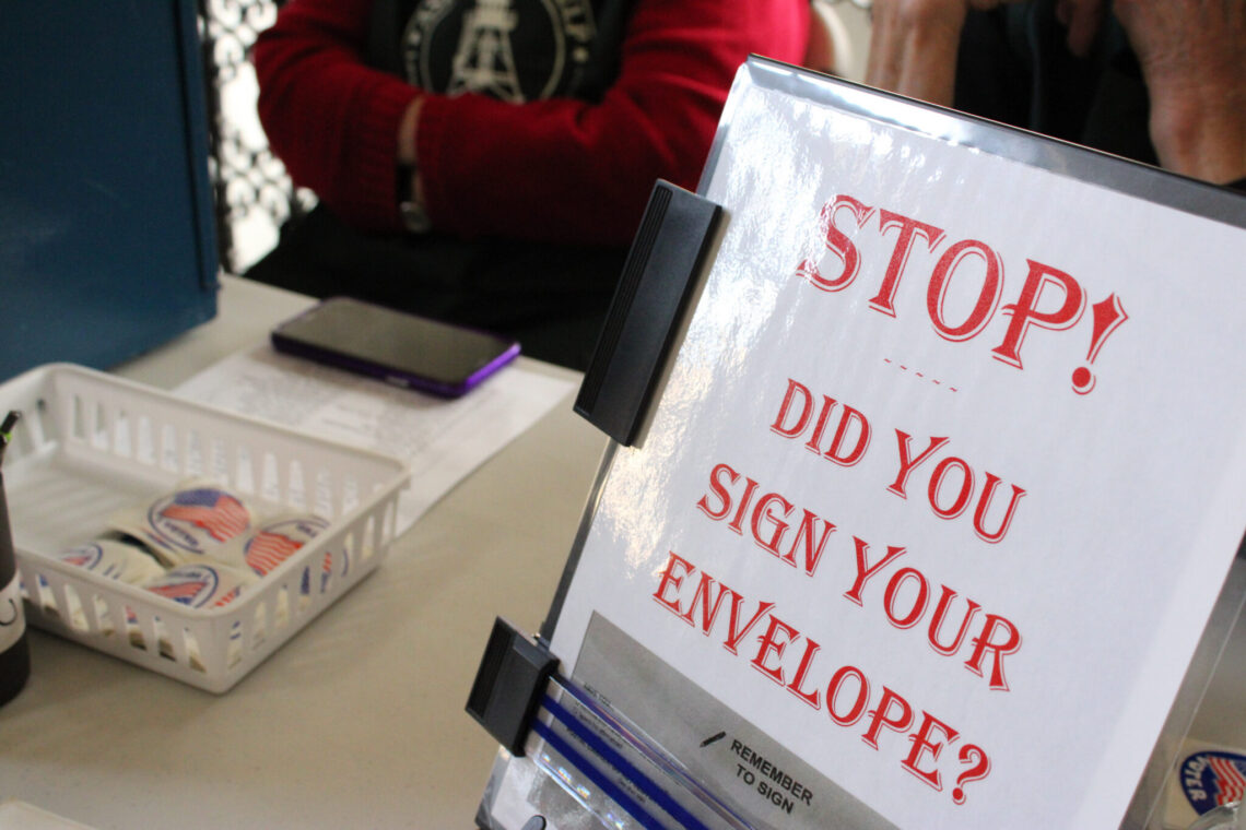 Signage on a desk asking: 'STOP! Did you sign your envelope?' with a binder and basket of stickers nearby.