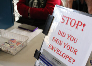 Signage on a desk asking: 'STOP! Did you sign your envelope?' with a binder and basket of stickers nearby.