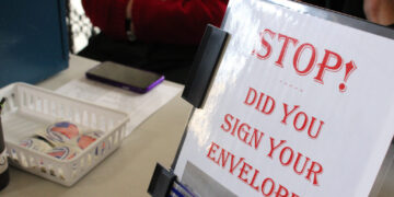 Signage on a desk asking: 'STOP! Did you sign your envelope?' with a binder and basket of stickers nearby.