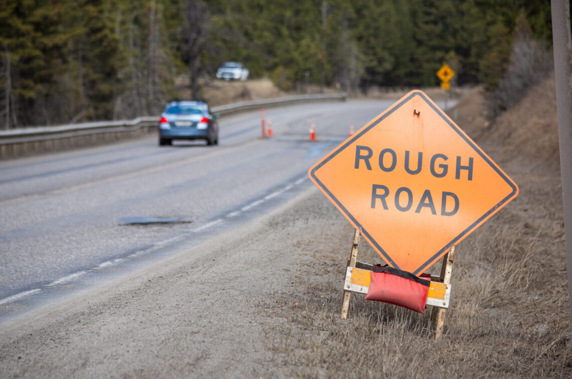 Orange diamond warning sign reading 'ROUGH ROAD' on a roadside near a winding, wet street with cars and orange cones in a forested area.