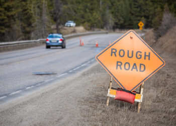 Orange diamond warning sign reading 'ROUGH ROAD' on a roadside near a winding, wet street with cars and orange cones in a forested area.