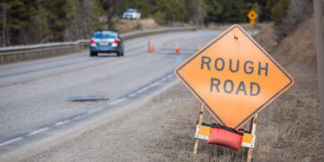 Orange diamond warning sign reading 'ROUGH ROAD' on a roadside near a winding, wet street with cars and orange cones in a forested area.