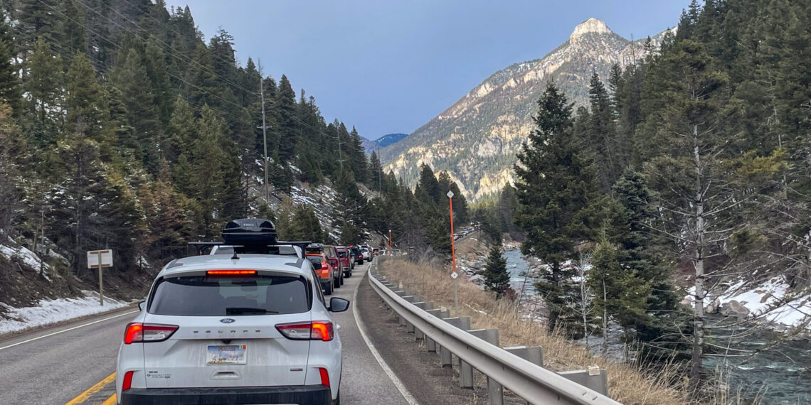 Line of cars in a mountain road traffic jam, with a metal guardrail on the right, evergreen trees, and a rocky peak in the distance under a blue sky.