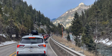Line of cars in a mountain road traffic jam, with a metal guardrail on the right, evergreen trees, and a rocky peak in the distance under a blue sky.