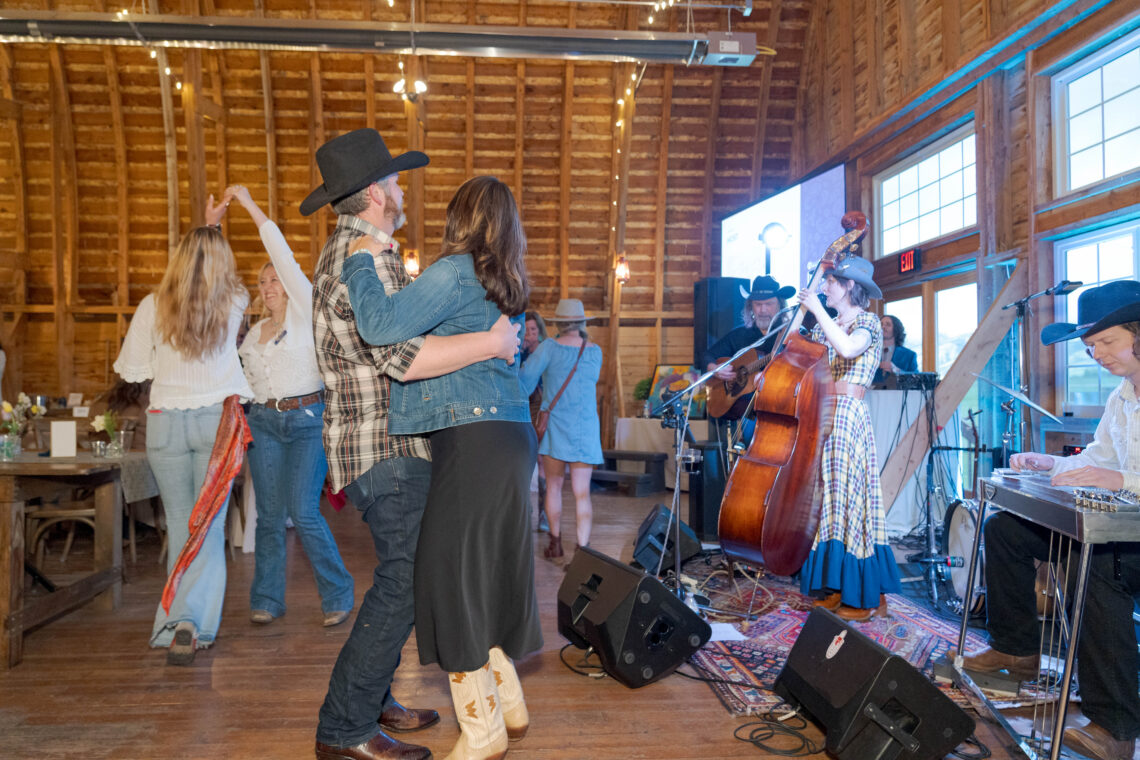 Couple dancing closely in a rustic wooden hall while a band plays nearby on stage.