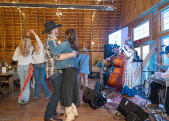 Couple dancing closely in a rustic wooden hall while a band plays nearby on stage.